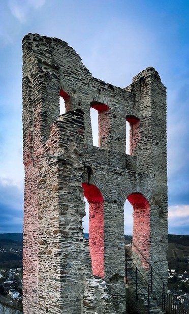 Gemäuerfragment der Ruine Grevenburg. Vor dämmrig blauem wolkenverhangenen Himmel steht die massive Wand mit Fenster- und Toröffnungen, deren Innenseite von einem roten Scheinwerfer außerhalb des Bildes angestrahlt werden 