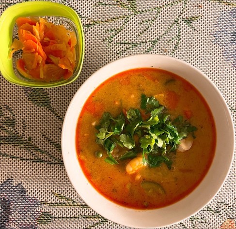 Auf einem Tisch mit einer Blumentischdecke stehen zwei Schalen: eine kleine grüne mit Kimchi und eine größere weiße mit der rotorangefarbenen Suppe, die mit Kräutern garniert ist