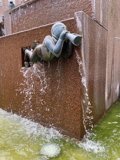 A sculpture of a figure playing a trumpet integrated into a fountain, with water streaming out from the trumpet and fountain walls.