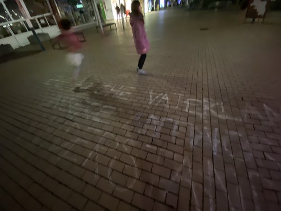 Zwei Kinder spielen nachts auf einem Ziegelsteinpflaster, mit undeutlichen Markierungen oder Spiegelungen auf dem Boden. Das Bild ist unscharf, was ein Gefühl der Bewegung vermittelt.