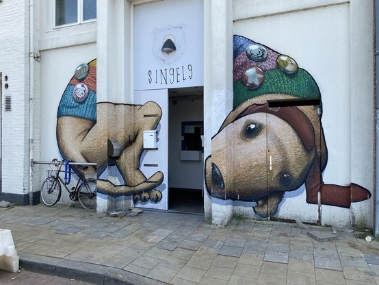 Street art depicting a dachshund in a beanie hat and a knitted sweater, its body is curling up with the building's doorway integrated as the split between the dog‘s head and body. A bicycle is parked to the left.