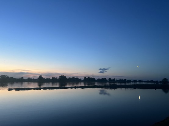 Dämmerungsszene mit einer Mondsichel, die sich auf dem ruhigen Wasser spiegelt, Silhouette von Bäumen am Horizont.