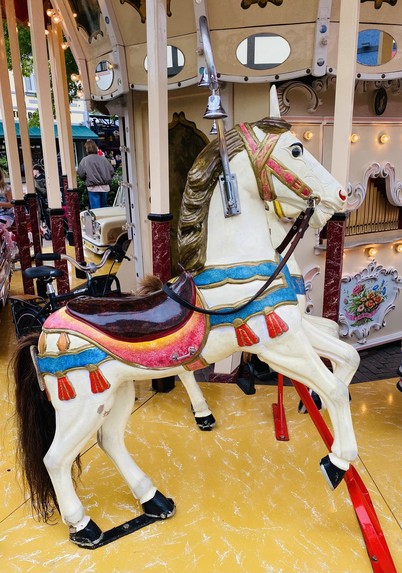 Carousel horse with elaborate decorations, part of a merry-go-round, with other rides and people in the background.