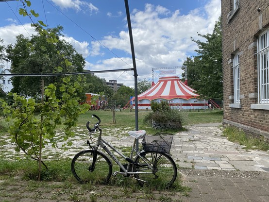Ein auf einem Kopfsteinpflasterweg abgestelltes Fahrrad mit einem rot-weiß gestreiften Zirkuszelt im Hintergrund, flankiert von Bäumen und einem Backsteingebäude mit vergitterten Fenstern auf der rechten Seite. Der Himmel ist bewölkt.