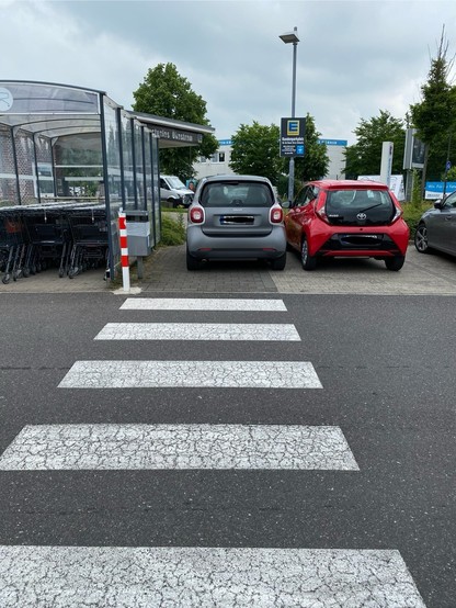 A pedestrian crossing on a parking lot of a supermarket with two cars parked across it, gray one is blocking the path. The red one is parked correctly. A shelter with shopping carts nearby are also visible.