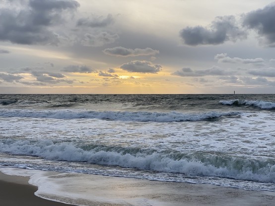 Sunset over the ocean with waves coming onto the shore and a cloudy sky.