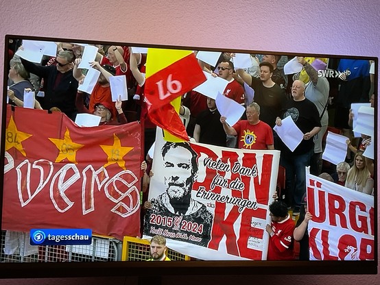 Eine Menge Fans, die weiße Papiere und Banner in einem Stadion halten. Banner zeigen Text und Bilder im Zusammenhang mit Fußball, darunter ein großes, das Jürgen Klopp für Erinnerungen von 2015 bis 2024 dankt.