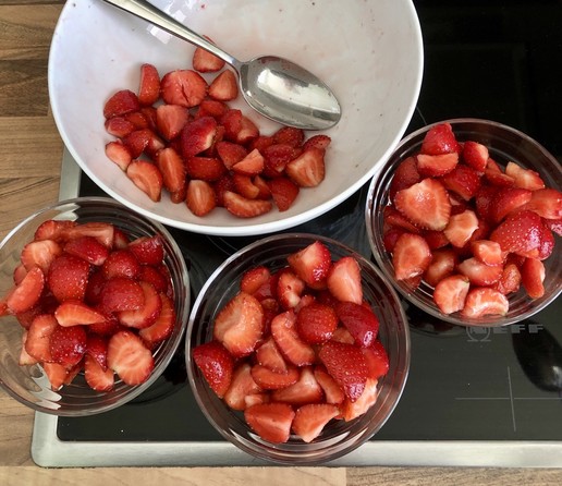 Eine größere weiße Schüssel mit ein paar geschnippelten Erdbeeren, daneben drei Schälchen voller Erdbeeren.