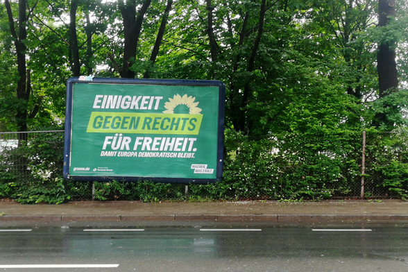 An einem Zaun am Rand einer Straße hängt ein großes Wahlwerbeplakat der Grünen mit ihrem Sonnenblumensymbol und dem Slogan 
