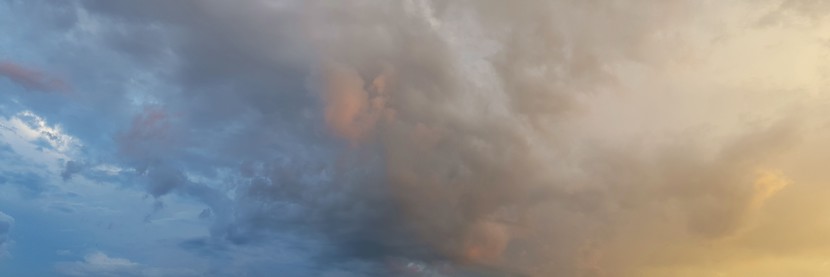 Panorama vom Himmel nach Gewitter: Links ist der Himmel blau bewegt bewölkt, von rechts schiebt sich eine Wolkenschicht heran, die von der rotgoldenen Abendsonne beschienen wird: mittig die Wolken leicht rötlich, rechts leuchtendes Grau mit Gelb, fast grünlich