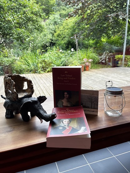 Eine Holzbank mit einem Keramik, Elefanten, einem Sonnenglas und den zwei Bänden der Übersetzung von Krieg und Frieden von Barbara Conrad. Dahinter ein Garten.