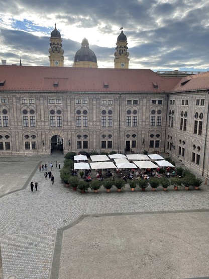 Blick aus dem Garderobentrakt des Münchner Herkulessaals, hoch oben unterm Dach, auf den Kaiserhof, bestückt mit Topfplanzen und Sonnenschirmen. Dahinter vor leicht bewölktem Abendhimmel die Türme der Theatinerkirche.