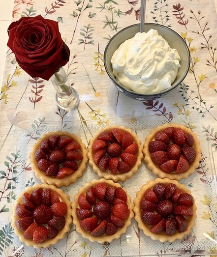 Sechs Tarteletts mit hübsch angeordneten halbierten Erdbeeren auf einer Glasplatte . Darüber ein Schüsselchen mit Schlagsahne und eine Vase mit einzelner Rose.
