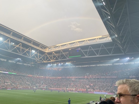 A crowded football stadium during a match with a rainbow visible above the open roof.