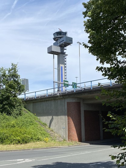 A tall control tower of an airport stands near an overpass, framed by trees and greenery under a partly cloudy sky.