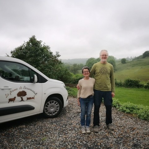 Ein freundlich lächelndes, handfestes älteres Paar steht vor grünen Hügel und neben einem weißen Auto mit dem Logo des B&B. 