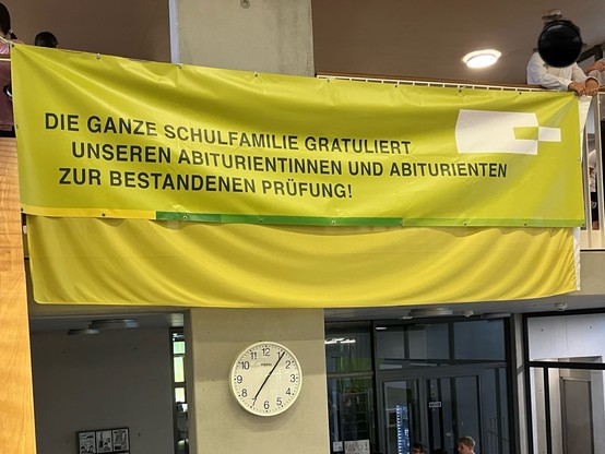 Ein gelbes Banner mit Text, der den Absolventen zum Bestehen ihrer Prüfungen gratuliert, hängt in einem Schulflur über einer Uhr. (autotext)