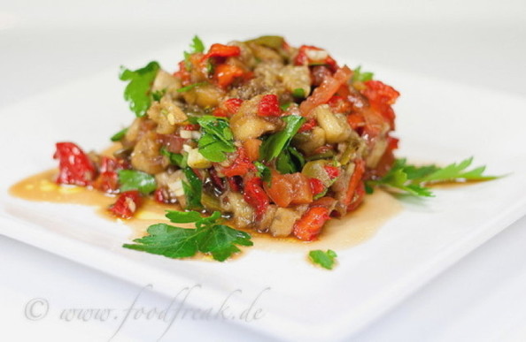 ein Salat aus gerösteten Auberginenwürfelchen, Tomaten und Paprika, mit Kräutern und Öl, zu einem Häufchen auf einem weißen Teller aufgeschichtet