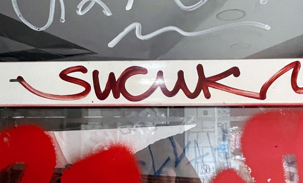 Nahaufnahme einer mit etlichen weißen und roten Graffiti beschriebenen und besprühten Glasfläche, im Fokus der dunkelrote, fette Markerschriftzug SUCUK