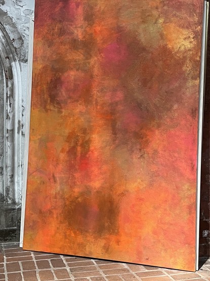 Ein sehr großes abstraktes Bild lehnt an einer Wand in der Lübecker Marienkirche. Das Bild zeigt eine farbige Fläche in Orange-Tönen. Von der Kirchenwand blättert Farbe, auf den Fußboden fällt ein Sonnenstrahl. 