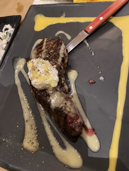 Gegrilltes Steak mit einem Klecks gewürzter Butter, mit Sauce beträufelt, auf einem schwarzen Teller mit einem Steakmesser mit Holzgriff serviert.