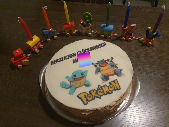 Geburtstagszug mit regenbogenfarbenen Kerzen und einer Acht, davor eine Torte mit einem Aufleger, auf dem zwei Pokemon abgebildet sind. 