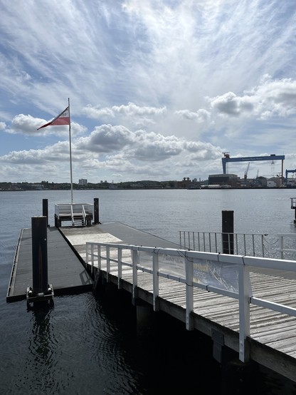 Blick auf die Kieler Förde mit Bootsanleger und Schleswig-Holstein-Flagge, auf den andern Uferseite der grosse HDW-Kran. Viel Himmel und Sonne und Wolken. 