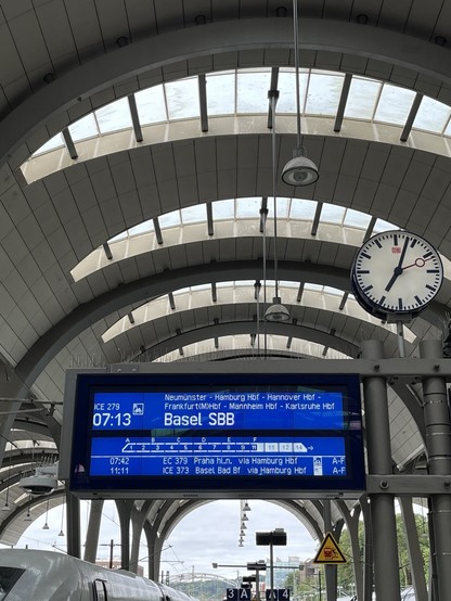 Das Abfahrtsgleis vom ICE nach Basel im Kieler Hauptbahnhof, mit Anzeigetafel, Bahnhofsuhr und gewölbtem Hallendach. 