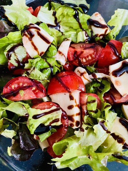 Tomaten-Mozarella-Salat mit Olivenöl, grobem Pfeffer, Balsamicocreme, Basilikum und Eichblattsalat. Angerichtet in eine Glasschale auf schwarzem Untergrund.