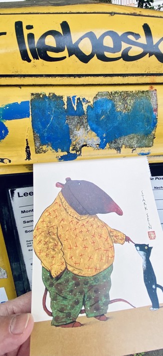 Eine Hand schiebt eine Postkarte in einen Postkasten, auf dem steht