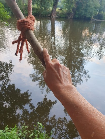 Eine Hand, die einen Stock hält, der an einem Seil befestigt ist. Auf dem zugehörigen Arm sind Wassertropfen. Im Hintergrund ist der Fluss und ein bisschen Grünzeug.