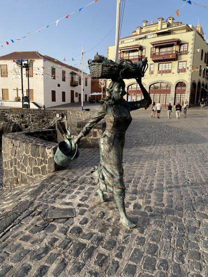 Statue of a woman carrying a basket on her head and holding a bucket with fishes, situated on a cobblestone street with buildings and colorful flags in the background. She is seemingly in a hurry.