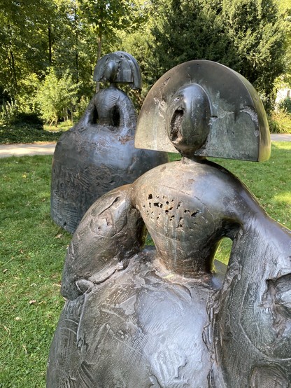 Two abstract bronze sculptures of figures with distinctive hairstyles, set against a green park background. The figures have rounded forms and detailed textures, evoking a sense of elegance and creativity.