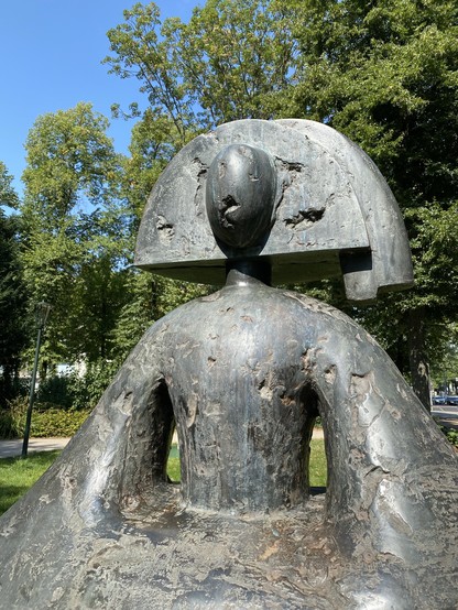 A bronze sculpture depicting a figure with a distinctive headpiece and a stylized body, set against a backdrop of green trees and blue sky. The artwork exhibits a rough texture and a minimalist design.