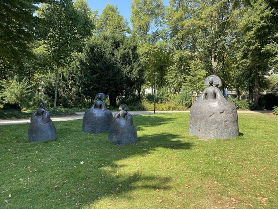 A group of four abstract sculptures positioned on green grass in a park, surrounded by trees. The sculptures feature rounded forms and heads, resembling human figures. Sunlight filters through the foliage, creating a tranquil outdoor setting.