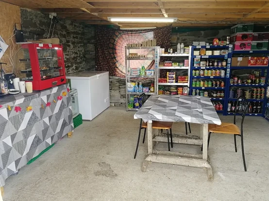 eine größere Garage mit einer Tiefkühltruhe und Lagerregalen, darin Lebensmittel in Packungen und Dosen, Reinigungsmittel usw. In der Mitte des Raums ein einfacher Tisch mit Stühlen, an einer Seite eine kleine Kühlvitrine, Kaffeeautomat und Becher