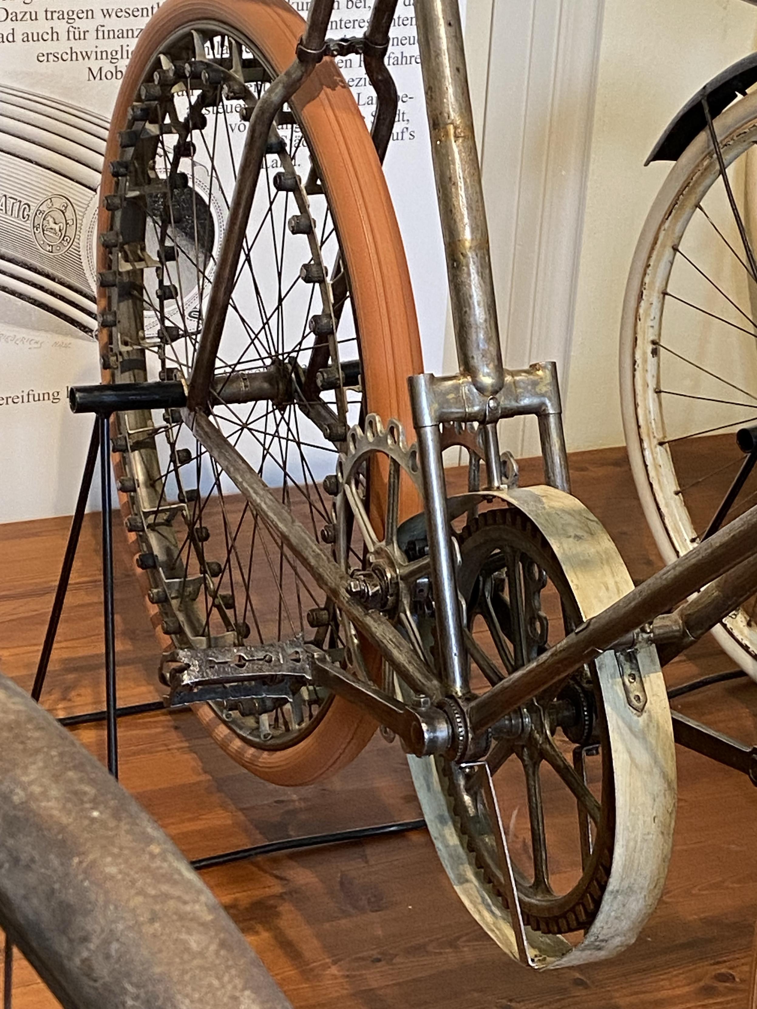 Eine Nahaufnahme des Hinterrads und des Antriebsstrangs eines Oldtimerfahrrads. Das Rad verfügt über einen einzigartigen orangefarbenen Reifen und der Metallrahmen zeigt Anzeichen von Verschleiß und Rost. Das Fahrrad wird auf einem Ständer ausgestellt, mit einem verschwommenen Hintergrund, der auf ein Museum oder eine Ausstellung hinweist
A close-up of the rear wheel and drivetrain of a vintage bicycle. The wheel features a unique orange tire, and the metal frame shows signs of wear and rust. The bicycle is displayed on a stand, with a blurred background indicating a museum or exhibit
Eine Nahaufnahme des Hinterrads und des Antriebsstrangs eines Oldtimerfahrrads. Das Rad verfügt über einen einzigartigen orangefarbenen Reifen und der Metallrahmen zeigt Anzeichen von Verschleiß und Rost. Das Fahrrad wird auf einem Ständer ausgestellt, mit einem verschwommenen Hintergrund, der auf ein Museum oder eine Ausstellung hinweist
A close-up of the rear wheel and drivetrain of a vintage bicycle. The wheel features a unique orange tire, and the metal frame shows signs of wear and rust. The bicycle is displayed on a stand, with a blurred background indicating a museum or exhibit