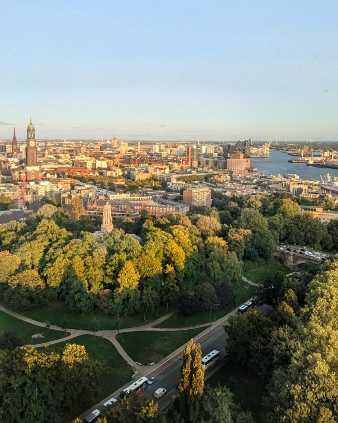 Blick von der Roof-Top Bar in den Tanzenden Türmen am Anfang der Reeperbahn in Richtung Osten. Es ist ein sehr milder Septembertag, 19:00 Uhr, kurz vor der blauen Stunde. Im Vordergrund ein Paar mit Denkmal. Das warme, stimmungsvolle Licht taucht die alte Innenstadt bis zum Hafen, rechts in ein warmes Licht.