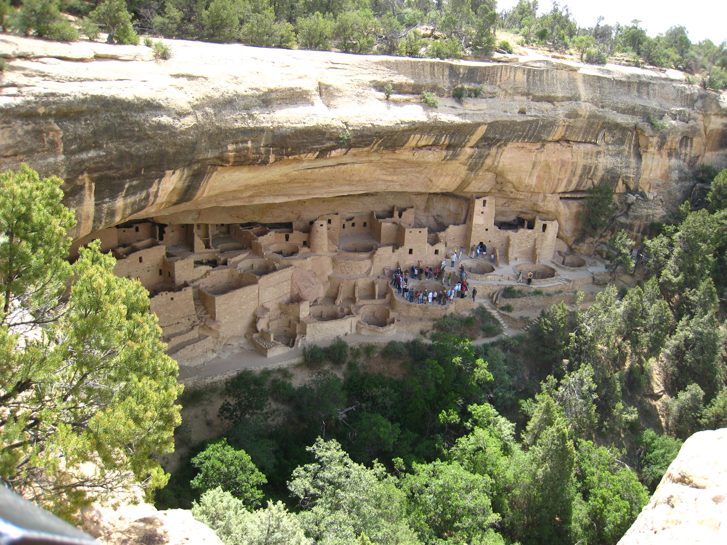In die Felsen eingebaute Wohnsiedlung "Cliff Palace". Es handelt sich um "gut erhaltene Felsbehausungen vorkolumbischer Anasazi-Stämme" (https://de.wikipedia.org/wiki/Mesa-Verde-Nationalpark). Im Vordergrund des Bildes ist ein bewaldeter, tieferer Grund zu sehen. Darüber erhebt sich ein Felsplateau in hellem Stein und senkrecht in dieses Plateau wurde eine Häusersiedlung gebaut, bei der der obere Teil des Plateaus wie eine Bedachung wirkt. In den Wänden der Behausungen sind Fensteröffnungen ausgespart, die den Blick ins Tal ermöglichen. Alle Häuser sind heute unbewohnt und eine Touristenattraktion.