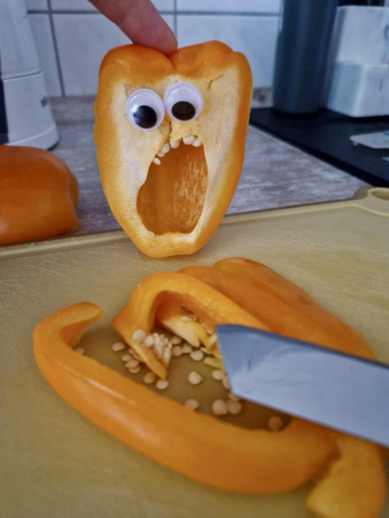 Eine halbierte gelbe Paprika mit Wackelaugen und einem überrascht-entsetzten „Gesichtsausdruck“ steht an einem Schneidebrett, während mehrere geschnittene Stücke Paprika auf dem Schneidebrett angeordnet sind. Ein Messer ragt ins Bild.