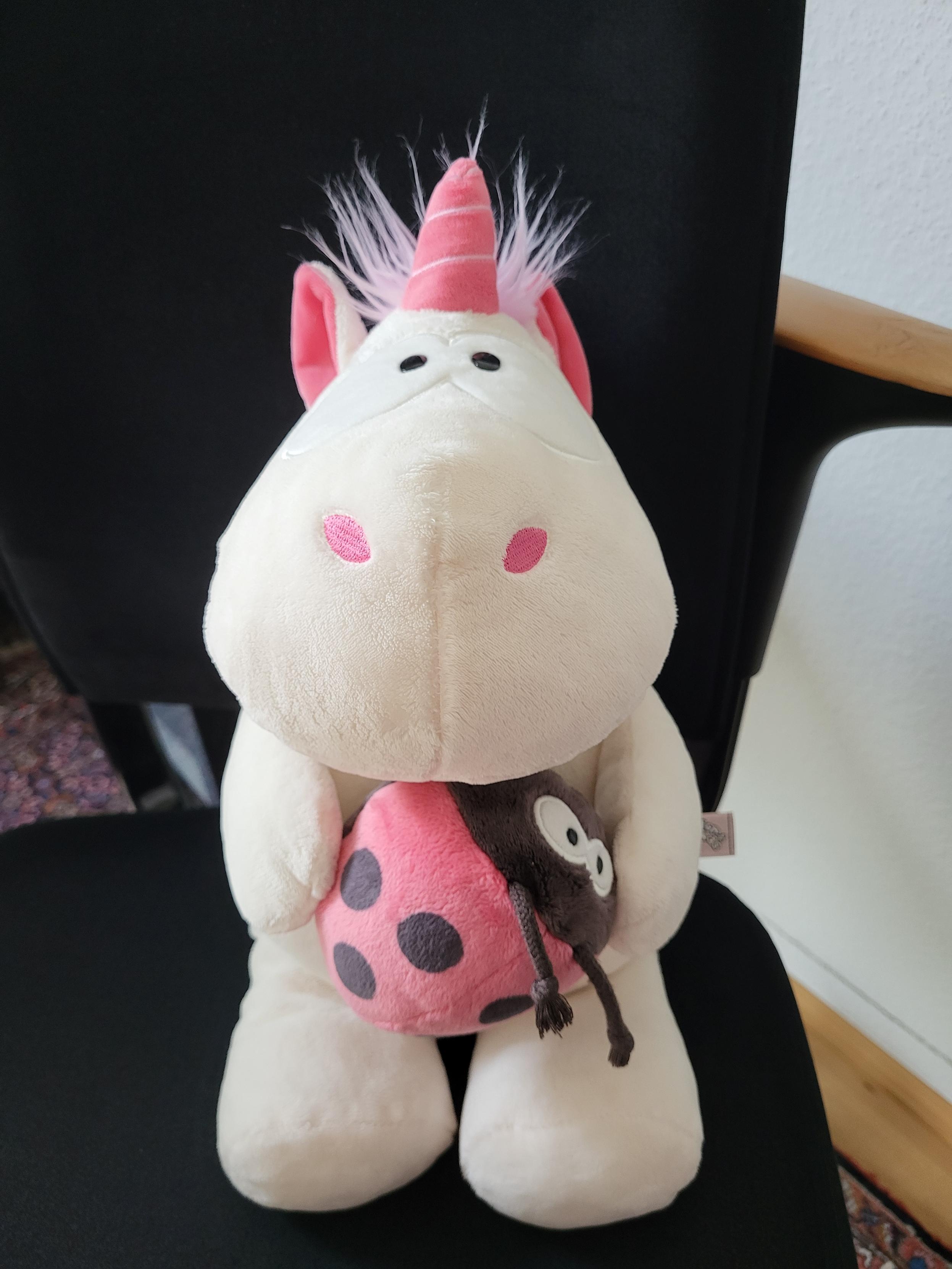 Ein weißes Stoffeinhorn mit rosa Mähne sitzt auf einem schwarzen Stuhl, es hat einen Stoff-Marienkäfer in den Armen.