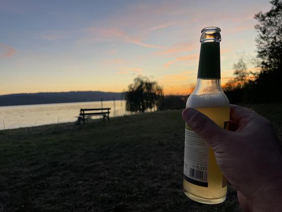 Ein Sonnenuntergang am Bodensee, Blick auf die Höri. An der rechten Seite ist meine Hand zu sehen, die ein Radler hält. 